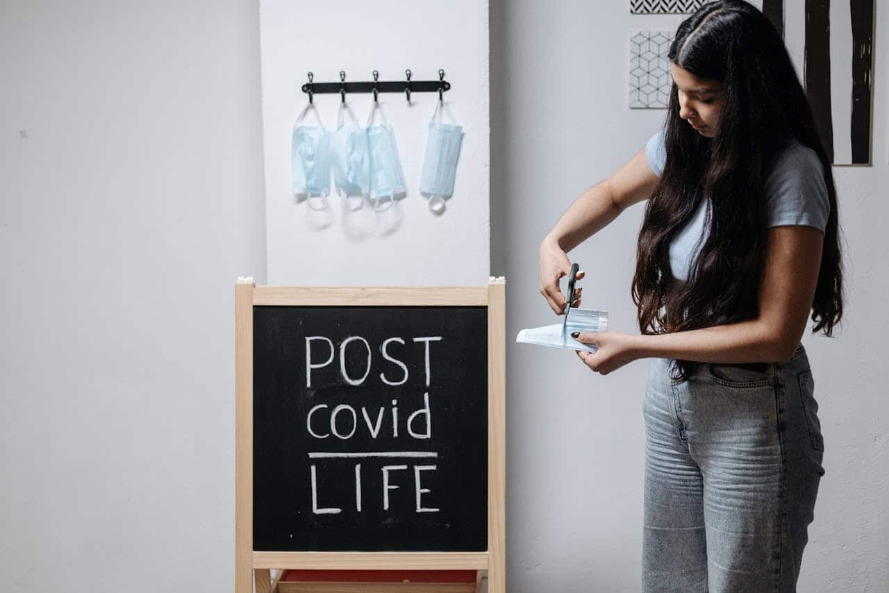 A young woman cutting a face mask next to a chalkboard reading POST covid LIFE indoors.